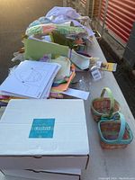 Wide view of a table showing assorted kids party and learning supplies including puzzles, coloring books, a terrarium planter kit, white apron, confetti box, and three small woven baskets.