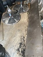 Top-down view showing the stainless steel shelf next to black chairs on a rough floor, highlighting surface scratches and wear on the shelf surface.