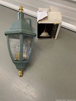 Greenish-gray outdoor hanging wall lantern with gold finials and two candle-style bulbs shown outside the box