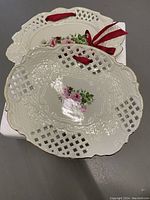 Top view of one porcelain wall hanging plate displaying pierced lattice edge, embossed details, and pink rose floral motif in center with red ribbon attached for hanging.