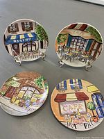 Four Avon decorative bistro themed plates arranged on gray surface, two on stands and two lying flat.
