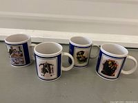 Four ceramic mugs arranged side by side showing different Norman Rockwell prints with a blue border on each mug