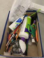 Box filled with paint brushes, scissors, adhesive bottles, pipe cleaners, and a clear decorative ornament with attached feather and paper