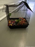 Full view of the black metal decorative bird cage with roof showing colorful artificial flowers lining the bottom tray.