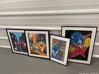 Four framed colorful prints displayed leaning against a wall: night cityscape, hot air balloons, abstract portrait, and multicolor figure