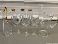 Front view of clear glassware including carafe with amber ribbon, three small corked bottles, three wine glasses, and a glass pitcher.