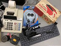 Photo showing a Sharp printing calculator with paper roll, a wireless keyboard, stereo headphones in packaging, a key finder or alarm device, and a household steamer box.