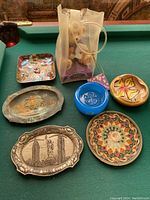 Seven small plates and trays arranged on a pool table with a plastic bag of dolls included.