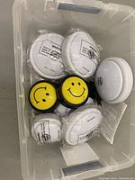 Top view of seven tap lights in a clear plastic bin showing four plain white and three yellow smiley face designs.