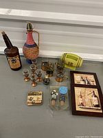 Full view of lot showing vintage bottles, six small copper-stemmed glasses, two larger glasses with copper bases and metallic lettering, green decorative glass bottle, coasters, and framed prints