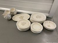 Photo showing vintage cream-colored porcelain plates, bowls, and cups with light brown floral motifs and pierced edges grouped together on floor.