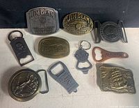 Ten metal belt buckles and keychains laid out on a white surface showing a variety of branded and themed designs including Jim Beam and fishing club.