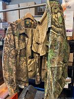 View of two Cabela's camouflage shirts and a beige fishing vest hanging together on a rack.