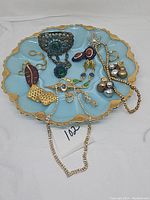 Overview of the blue glass egg-shaped plate containing assorted costume jewelry including brooches, earrings, necklace, and pins.