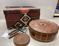 Photo showing carpeted storage box, round wooden bin with lid, and a wooden cutting board with metal handle along with metal kitchen utensils with wooden handles