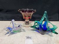 Photo showing all three glass pieces together: purple footed bowl, blue-green sculptural centerpiece, and smaller basket with handle and Murano sticker