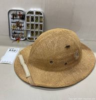 Photo showing the Wesco woven straw fishing hat in front with Cabela’s aluminum fly fishing lure box open displaying various flies and lures in the background.