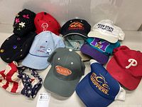 Photo of eleven vintage hats laid out showing various logos including Phillies, Orange County Choppers, Von Dutch, and Penn State.