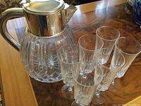 Photo showing a 24% lead crystal jug with silver plate lid and handle, along with six hand cut crystal goblets arranged on a wooden table.
