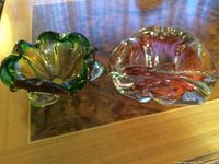 Two Murano glass bowls placed on a wooden surface with a clear glass panel, showing the color, shape, and overall condition of the items.