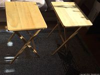 Two solid wood TV tables with foldable X-shaped legs photographed in natural light on carpeted floor. Light wood finish and rectangular tops visible.