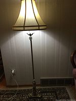 Photo of a brass floor lamp with a beige fabric lampshade turned on, casting warm light on a patterned carpet and wallpapered wall.