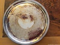 Top view of the round silver plate tray showing intricate engraved swirl pattern on the tray face and the raised beaded edge. The tray shows some light surface tarnish and reflections.