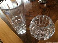 Photo showing two clear lead crystal vases on wooden surface: one taller rectangular vase, square-shaped with vertical and horizontal facets forming a grid. Second smaller round vase with similar grid cut pattern and starburst on base.
