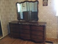 Long dresser with 9 drawers in walnut veneer, topped by a large decorative framed mirror, positioned against wall with vintage wallpaper.