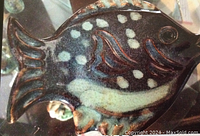 Close-up image of a handmade pottery fish sculpture with dark brown glaze, white spots, and carved fin details.