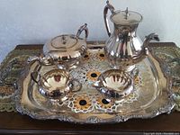 Photo showing full silver coffee and tea service set on lace doily atop a decorative table cloth. Includes large tray, coffee pot, teapot, creamer, and sugar bowl.
