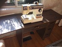 Singer sewing machine set on a wooden table with drawers and foldable extension wings, visible in natural light near a window.