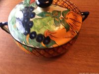 Close-up of the stoneware bowl with lid showing bright hand-painted fruit and leaf pattern with a black knob on the lid.