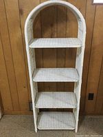 Photo of a white wicker shelving unit with four shelves, approximately 6 feet tall, placed against a wooden wall.