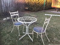 Outdoor image of vintage white wrought iron cafe-style table frame and two matching chairs with black seats, set on grass.