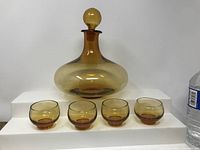 Vintage amber glass liquor decanter with round stopper and four matching amber glass shot glasses, shown on stepped white platforms.