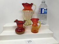 Three vintage glass pitchers including large cranberry pitcher with eagle design, and two smaller amberina style pitchers, all placed on white steps next to a water bottle for scale.