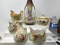 Full grouping of 6 porcelain decorative items including vases, pitchers, and a sugar pot displayed on white risers with water bottle for scale