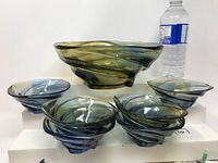 Seven art glass bowls comprising one large bowl and six small bowls, displayed with a water bottle for scale. Colors include green, blue, and yellow swirls.