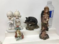Four figurines: two boy and girl child figurines in white, black boar on a base, rustic angel figurine with wings and lantern behind.