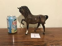 Side view of large glossy dark brown horse figurine with white hoof and swishing tail, placed next to a soda can for scale.