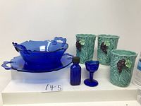 Seven items displayed: cobalt blue two-handled glass bowl and matching server plate, cobalt blue eye wash glass, cobalt blue medicine bottle, and three green Napoware cups with grape and leaves design.