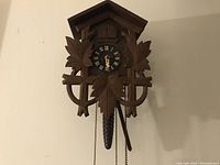 Full front view of carved wooden cuckoo clock hanging on wall, showing leaf decorations, clock face and hanging pine cone weight.