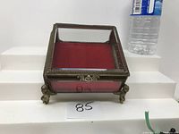 Front view of glass jewelry box showing red velvet lining, cherub-shaped feet, and metal frame with ornate detailing.