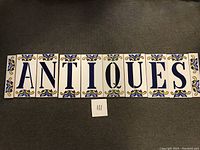 Full set of 10 Italian ceramic tiles arranged to spell 'ANTIQUES' showing 8 letter tiles and 2 end tiles with floral decorations.