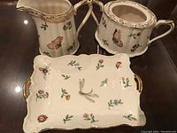 Photo of creamer, lidded sugar bowl, and serving tray showing detailed nature-themed designs and gold trim.
