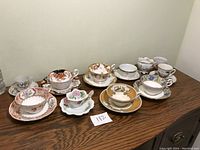 The full lot of 11 cups and 9 saucers arranged on a wooden surface showing variety in floral and decorative patterns with gold trim.