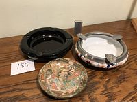 Three vintage ashtrays on wooden surface showing black ceramic, decorated white and floral, and detailed printed metal ashtray