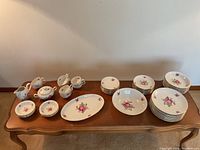 Full view of the partial china set arranged on a table including cups, saucers, plates, small bowls, and serving platter, all featuring Vienna Rose floral design with pink roses and gold trim.