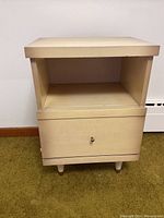 Front view of beige wood cabinet showing open shelf and drawer with knob, standing on four short legs.